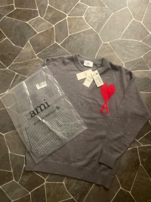 Grå stickad tröja från Ami med rött hjärta - Säljer en grå stickad tröja från Ami Alexandre Mattiussi med ett stort rött hjärta och bokstaven A broderat på bröstet. Tröjan har rund hals, ribbade muddar och lång ärm. Perfekt för dig som gillar stilrena detaljer och vill sticka ut med en ikonisk design.