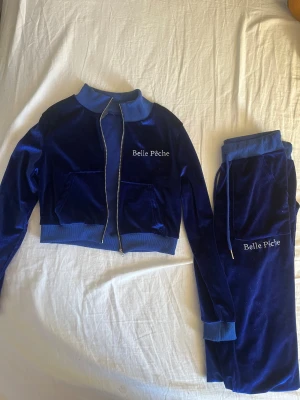 Blått mjukis set - Mörkblå mjukisbyxori mjuk velour från Belle Pêche. Byxorna har bootcut-modell, resår och snörning i midjan samt broderad logga på ena benet. Matchande jacka med dragkedja och ribbade muddar ingår. 