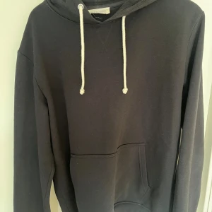 Svart hoodie från Pier One - Svart hoodie från Pier One med klassisk huva och vita snören. Tröjan har en stor magficka framtill och långa ärmar. Perfekt för en chill och avslappnad stil.