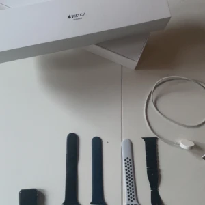 Apple Watch Series 3 med flera armband - Apple Watch Series 3 med svart fyrkantig urtavla och flera utbytbara armband: svart silikon, svart metall och vitt sportarmband med svarta hål. Laddkabel och originalförpackning ingår. Perfekt för dig som vill ha en smart och stilren klocka. Jag har även några stretch armband de är bara o skriva om du är intresserad.