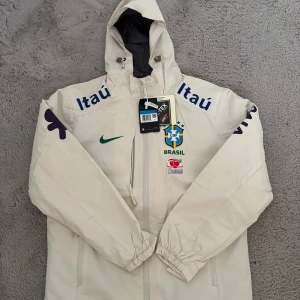 Nike Brasil CBF Gore-Tex vindjacka - Exklusiv Brasil CBF vindjacka från Nike i ljusbeige med blå och gröna detaljer. Jackan har huva, dragkedja, flera loggor och är tillverkad i Gore-Tex-material med Dri-Fit-teknologi. Perfekt för dig som vill ha sportig stil och skydd mot väder.