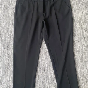 Svarta kostymbyxor från Cambio - Svarta kostymbyxor från Cambio med rak passform och elastisk midja. Byxorna har klassiska fickor fram och bak, bälteshällor och stängs med knapp och dragkedja. Snyggt stilrena och passar perfekt till en dressad look. Mått Midjan: ca 47 cm Längd från grenen och ner: ca 74 cm  Kommer från ett djur och rökfritt hem.