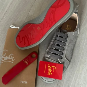 Louboutin - Snygga grå sneakers från Christian Louboutin i mjuk mocka med klassisk rund tå och platt sula. Skorna har mörkgrå snörning och den ikoniska röda undersulan med Louboutin-logga. Perfekt för dig som vill sticka ut med exklusiv stil. Helt oanvända med prislapp!