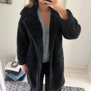 Svart teddyjacka med stora knappar - Mysig svart teddyjacka med stora slag och två knappar framtill. Jackan är lång, har bred krage och är helfodrad för extra värme. Perfekt för dig som gillar en oversized och chill look. Passar till allt och är superenkel att styla.