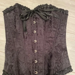 Svart korsett med snörning och rosett - Säljer en helt ny svart korsett med snyggt mönstrad yta och glansiga detaljer. Den har snörning både fram och bak, dekorativa rosetter och volangkant upptill och nedtill. Perfekt för dig som vill ha en edgy och elegant look med extra attityd. Perfekt till halloween 