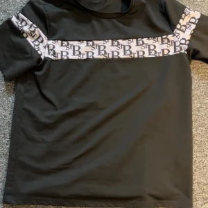 Svart t-shirt med logoband - Svart t-shirt från SikSilk med ett brett, ljust band över bröstet och ärmarna där loggan upprepas i svart. Klassisk rund hals och rak passform. Perfekt för dig som gillar streetwear och vill sticka ut med snygga detaljer.
