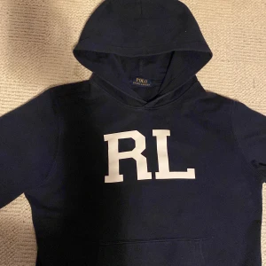 Mörkblå hoodie Polo Ralph Lauren - Snygg mörkblå hoodie från Polo Ralph Lauren med stor vit RL-logga på bröstet. Klassisk huva och känguruficka framtill. Perfekt för dig som gillar stilrena och sportiga plagg med premiumkänsla.