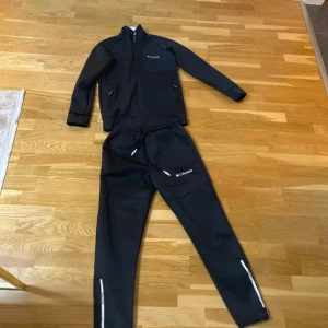 Svarta mjukisbyxor från Columbia - Svarta mjukisbyxor från Columbia med vit logga på benet och dragkedjor vid bensluten. Byxorna har snörning i midjan och är tillverkade i ett mjukt syntetmaterial. Perfekta för chill eller träning, med en clean och sportig look.