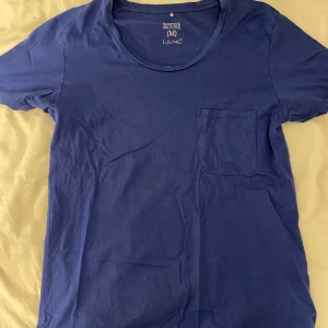 Blå t-shirt med ficka från Ljung - Snygg blå t-shirt från Ljung i storlek M, tillverkad i 100% bomull. Modellen har en scoopneck och en praktisk bröstficka på vänster sida. Perfekt för dig som gillar enkel och stilren design med en modern touch.