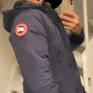 Mörkblå dunjacka från Canada Goose - Säljer en mörkblå Canada Goose dunjacka med huva och ikonisk patch på ärmen. Jackan har dubbelknäppning med både dragkedja och knappar, två stora fickor framtill och ribbade muddar vid ärmslut. Perfekt för kalla dagar och riktigt snygg stil. Har mycket mer bilder på den om du är intresserad, priset går och diskutera 