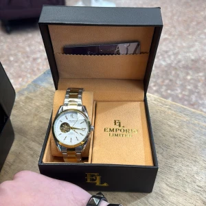 Emporio Limited automatisk herrklocka - Snygg automatisk klocka från Emporio Limited med tvåfärgad länk i silver och guld. Urtavlan är vit med gulddetaljer och har ett öppet fönster som visar mekanismen. Klockan levereras i original ask med logga och exklusivt medlemskort. Automat 
