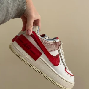 Nike Air Force 1 Shadow vit/röd - Nike Air Force 1 Shadow sneakers i vit och röd med dubbla detaljer och chunky platt sula. Skorna har snörning, perforerad tå och röd swoosh på sidorna. Ovandelen är i skinn och syntet, med ljusgrå meshfoder och unika lager på häl och sula.