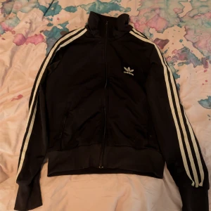 Svart Adidas track jacket med vita ränder - Säljer en svart Adidas trackjacket med klassiska vita ränder längs ärmarna och stort Adidas-tryck på ryggen. Jackan har hög krage, dragkedja framtill och ribbade muddar. Perfekt för en sportig och chill stil.