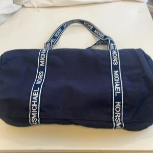 Blå duffelbag från Michael Kors - Snygg marinblå duffelbag från Michael Kors med vita logoband och silverfärgade dragkedjor. Väskan är cylinderformad och har två handtag, perfekt för gymmet eller weekendresan. Tillverkad i slitstark polyester med tydlig logga på banden.