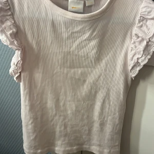 Ljusrosa ribbad topp med volangärm Gina Tricot - Supersöt ljusrosa ribbad topp från Gina Tricot med korta volangärmar i broderad bomull. Toppen har rund halsringning och är i mjukt material, perfekt för dig som gillar romantiska detaljer och en feminin look.