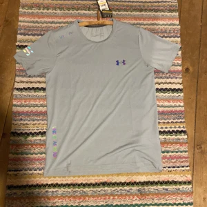 Ljusgrå Under Armour HeatGear t-shirt - Snygg ljusgrå t-shirt från Under Armour med färgglatt UA-logga på bröstet och tryck vid axeln. Modellen är kortärmad och har rund hals. Materialet är polyester och designen är perfekt för träning. Diskret text 'UA-SPEC' och 'Live Fear You' på sidan.