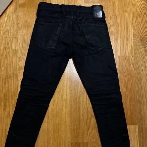 Svarta Replay Anbass jeans, W30 L32 - Säljer ett par svarta Replay Anbass jeans med slim passform och klassisk femficksdesign. Jeansen har svarta sömmar, Replay-logga på bakfickan och läderpatch i midjan. Materialet är stretchigt jeans för skön komfort och snygg siluett.
