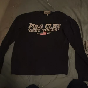 Mörkblå  tröja Saint Vincent - Svart långärmad tröja från Saint Vincent med stor vit text 'Polo Club Saint Vincent' och amerikansk flagga framtill. Klassisk crewneck-modell, perfekt för en sportig och avslappnad look. Skön passform och stilren design.