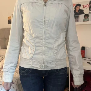 Vit jacka - Snygg vit vindjacka med dragkedja framtill och två fickor. Jackan har ribbade muddar vid ärmslut och nederkant samt dekorativa knappar vid midjan. Perfekt för dig som gillar en clean och sportig stil.