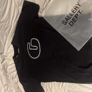 Svart t-shirt från Gallery Dept. - Svart t-shirt från Gallery Dept. med stor vit logga framtill och tryck i rosa och grått på ryggen. Klassisk rund hals och korta ärmar. Materialet är mjuk bomull, perfekt för dig som gillar streetwear med coola prints.