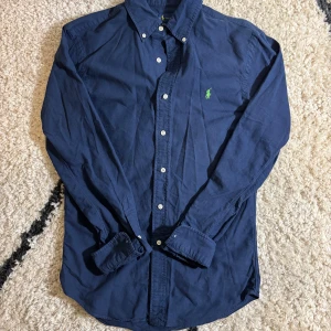 Ralph Lauren Skjorta - Säljer denna snygga marinblå skjorta från Ralph Lauren i slim fit-modell. Skjortan har en snygg neongrön logga på brösten. Skjortan är använd sparsamt och saknar defekter. Modellen är 181cm och väger 72kg.  Tveka inte vid frågor eller prisförslag🤝✍️!