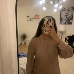 Beige blus med veckade ärmar - Säljer en beige blus med långärmade, lätt puffade ärmar och knappar vid ärmsluten. Blusen har en högre halsringning med fina veckdetaljer framtill. Perfekt för dig som gillar en stilren och chic look.