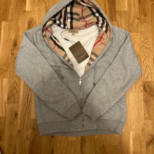 Grå Burberry hoodie med rutigt foder  - Snygg grå hoodie från Burberry med dragkedja och klassiskt rutigt foder i huvan. Tröjan har snörning vid huvan och ribbade muddar. Perfekt för dig som vill ha en stilren och lyxig look med ikoniska Burberry-detaljer.