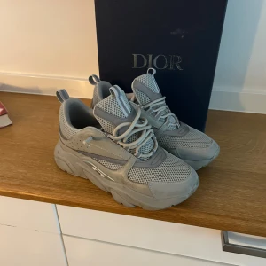 Grå chunky sneakers från Dior - Säljer ett par grå chunky sneakers från Dior med mesh och syntetdetaljer. Skorna har en riktigt fet sula, snörning och coola paneler i olika grå nyanser. Perfekta för dig som vill sticka ut med exklusiva och trendiga sneakers.