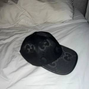 Svart keps från Gucci med GG-mönster - Säljer en svart keps från Gucci med ikoniskt GG-mönster över hela kepsen. Kepsen har böjd skärm och justerbar rem baktill. Tillverkad i slitstarkt tyg med detaljer i svart. Perfekt för dig som vill ha en lyxig och stilren accessoar.
