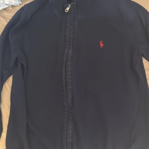 Marinblå kofta från Ralph Lauren - Snygg marinblå kofta från Ralph Lauren med dragkedja framtill och klassisk röd broderad logga på bröstet. Tröjan har ribbad krage och är långärmad, perfekt för lager-på-lager-stil. Passar dig som gillar stilrena och tidlösa plagg.