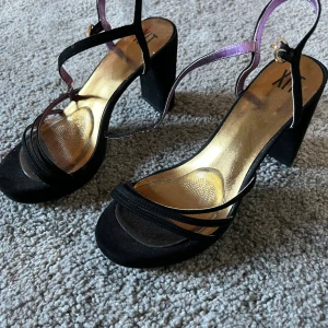 Svarta sandaler med blockklack från XIT - Snygga svarta sandaler från XIT med bred blockklack och tunna remmar i mocka. Insidan är guldig och remmarna har en lila detalj samt spänne i guld. 