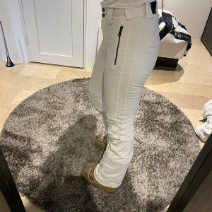 Vita skidbyxor från J.Lindeberg - Snygga vita täckbyxor från J.Lindeberg med dragkedjor framtill och markerad midja. Byxorna har bootcut-snitt och är perfekta för vintern. De är tillverkade i ett slitstarkt material och har bälteshällor samt detaljerade sömmar för extra stilpoäng.Nypris 3700kr och köpta i butik, del har blivit använda och tvättade, men de har ett par års användning kvar i sig sista bilden visar byxorna men det syns ej när dem är på utan bara när man tagit av sig dem 