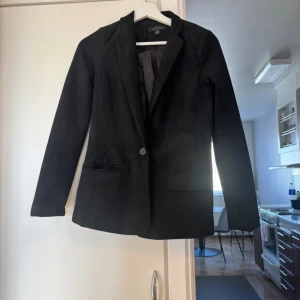 Svart kavaj från Amisu - Stilren svart kavaj från Amisu med klassisk passform och en knapp framtill. Kavajen har två fickor med lock och är helfodrad. Perfekt för dig som vill ha en enkel och snygg look.