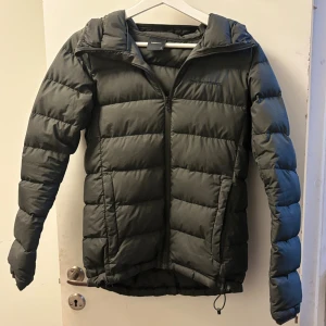 Grön grå dunjacka från Peak Performance - Säljer en grön dunjacka från Peak Performance med klassisk pufferstil. Jackan har dragkedja framtill, hög krage och två sidofickor. Perfekt för kalla dagar och har en stilren look som funkar till det mesta.