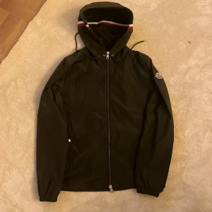 Svart vindjacka från Moncler med huva - Svart vindjacka från Moncler med hel dragkedja, huva med röd och vit rand, samt Moncler-logga på ärmen. Jackan har dragsko i huvan och två sidofickor. Perfekt för dig som vill ha en stilren och sportig look.