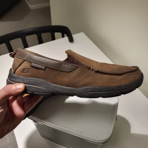Bruna loafers från Skechers, storlek 42 stora i Storlek  - Säljer ett par bruna loafers från Skechers i storlek 42. Skorna har en snygg, rund tå och är tillverkade i mocka och syntet med robust svart sula. De har en enkel slip-on design utan snörning och diskreta sömmar som ger en chill look.