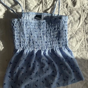 Blommig blå linnetopp från Gina Tricot - Supersöt ljusblå linnetopp från Gina Tricot med smockad överdel och tunna axelband. Toppen har små vita och mörkblå blommor och en lätt volang nertill. Perfekt för varma dagar och ger en fräsch sommarkänsla.