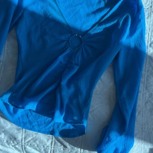 Blå långärmad topp från Gina Tricot - Säljer en blå långärmad topp från Gina Tricot i storlek XS. Toppen har v-ringning med en snygg ringdetalj framtill och är gjord i ett lätt, strukturerat syntetmaterial. Perfekt för dig som vill sticka ut med en färgstark look.