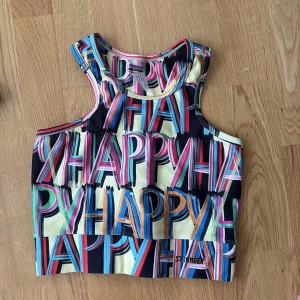 Färgglad träningstopp och matchande byxorfrån Stronger XS - Säljer en sportig och färgglad linnetopp och träningsbyxor från Stronger i storlek XS. Toppen har ett grafiskt mönster med ordet 'HAPPY' i flera färger som blå, rosa, grön, gul och orange. Materialet är stretchigt och består av 85% återvunnen polyester och 15% elastan.