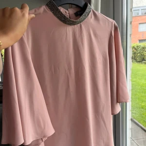 Ljusrosa klänning med glittrig krage - Supersöt ljusrosa klänning med glittrig krage och vida, korta ärmar. Klänningen har en rak passform och är tillverkad i ett lätt, flowy material som ger en elegant känsla. Perfekt för dig som vill sticka ut med en touch av glitter.