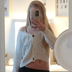 Vit croppad kofta med knappar - Säljer en vit croppad kofta med stora mörka knappar och v-ringning. Koftan är stickad i ett mjukt material och har lång ärm. Snygg att bära offshoulder för en chill vibe. Perfekt till jeans eller kjol.