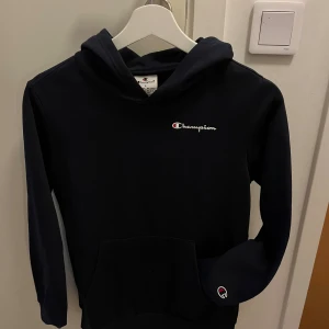 Marinblå hoodie från Champion L - Snygg marinblå hoodie från Champion i storlek L/11-12 år. Klassisk design med broderad Champion-logga på bröstet och ärmen. Tröjan har huva och en stor magficka framtill. 