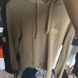 Beige hoodie från Morris med tryck - Snygg beige hoodie från Morris med broderat emblem och text på bröstet. Tröjan har huva med snörning och långa ärmar. Tillverkad i mjuk bomull som är skön att ha på sig. Perfekt för dig som gillar stilrena och klassiska plagg.