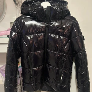 Svart glansig pufferjacka från Khujo - Säljer en svart glansig pufferjacka från Khujo med huva och dragkedja framtill. Jackan har två fickor med dragkedja och coola detaljer som logotyp på ärmen och huvan. Använd en säsong och den har inga defekter! Jag har vanligtvis storlek xs/s och denna sitter perfekt! Nypris 2400kr