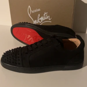 Christian Louboutin svarta sneakers med nitar - Säljer ett par svarta sneakers från Christian Louboutin med ikonisk röd sula och svarta nitar på tån. Skorna har snörning och är tillverkade i skinn och mocka för en lyxig känsla. Kommer med originalpåse och äkthetsbevis.