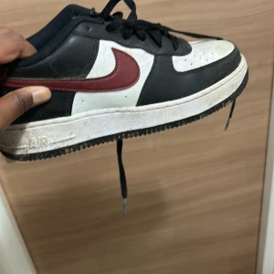 Nike Air Force 1 svart/vit/bordeaux - Säljer ett par Nike Air Force 1 sneakers i svart och vitt med bordeauxröd swoosh och häl. Klassisk låg modell med snörning, rund tå och platt sula. Ovandel i skinn och textil, perfekt för dig som gillar streetstyle och tidlös design.