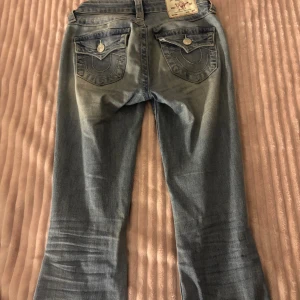 True Religion Becca Bootcut Jeans - Snygga ljusblå bootcut jeans från True Religion, modell Becca Mid Rise Bootcut. Klassiska fickor med dekorativa sömmar och knappar bak. Jeansen är i denim med slitningar och fransade detaljer vid benslut. Perfekt för dig som gillar en avslappnad och trendig look.
