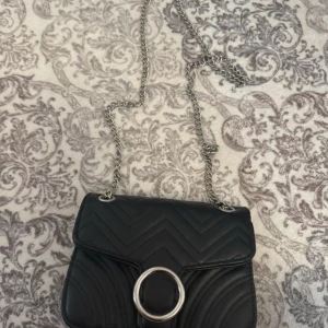 Svart quiltad axelväska med kedja - Snygg svart axelväska i quiltat mönster med silvrig kedjerem och stor rund metallring framtill. Väskan har en kompakt form och är tillverkad i skinnimitation. Perfekt för dig som vill ha en stilren och trendig accessoar.