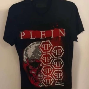 Svart t-shirt från Philipp Plein med dödskalle, T-shirten har tappat lite stenar som knappt syns i övrigt är den i nyskick - Cool svart t-shirt från Philipp Plein med stor dödskalle i rött, silver och vitt framtill. T-shirten har tryck med loggor och grafiska detaljer, samt korta ärmar och rund hals. Perfekt för dig som gillar edgy streetstyle.
