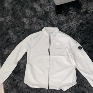 Vit vindjacka från C.P. Company - Snygg beige overshirt från C.P. Company med svart dragkedja och klassisk linsdetalj på ärmen. Jackan har en stilren design med bröstficka och stårkrage, perfekt för dig som gillar streetwear och cleana looks.